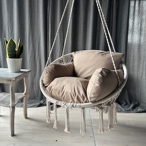 May include: A white macrame hanging chair with a beige cushion and two pillows. The chair is hanging from the ceiling and is in front of a gray curtain.