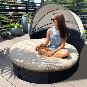 Op de afbeelding: Een ronde buitendagbed met een zwarte rotan basis en een beige kussen. De daybed heeft een luifel en staat op een houten terras. Op de achtergrond staan ​​verschillende potplanten.