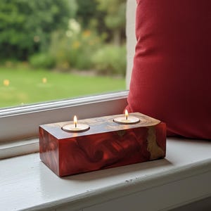 May include: A rectangular, red-toned candle holder with two lit tea light candles. The holder has a swirled, marbled appearance and sits on a white windowsill. A red pillow is in the background, with a view of a green lawn and trees outside the window.