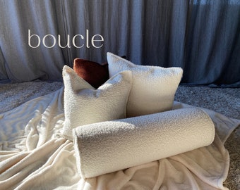 May include: A collection of soft, textured pillows and a bolster in cream and rust colors. The word "boucle" is displayed in the upper left corner. The pillows are arranged on a cream-colored blanket.