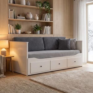 Puede incluir: Un sofá cama blanco con tres cajones, cojines grises y almohadas. El sofá cama está en una habitación con estanterías de madera, libros, plantas y una chimenea. Dos alfombras de color neutro están en el suelo de madera.