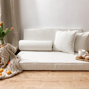 May include: A cozy, off-white pet bed with a textured surface, featuring a bolster pillow and two square pillows. A wooden tray with muffins and a cup sits nearby. A knitted blanket and a vase of orange tulips add warmth.