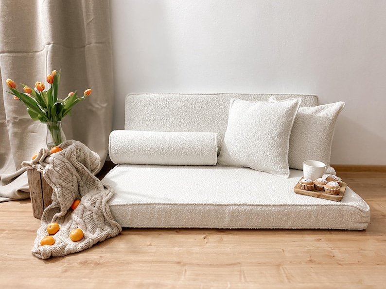 May include: A cozy, off-white pet bed with a textured surface, featuring a bolster pillow and two square pillows. A wooden tray with muffins and a cup sits nearby. A knitted blanket and a vase of orange tulips add warmth.