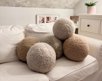 May include: Five round, fluffy, beige and cream colored throw pillows on a white couch. The pillows are arranged in a pyramid shape.