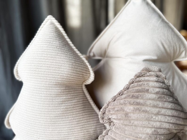 May include: Three decorative tree-shaped pillows in varying shades of white and beige. The front pillow has a ribbed texture, while the others have a smooth, soft appearance. These pillows are designed for home decor.
