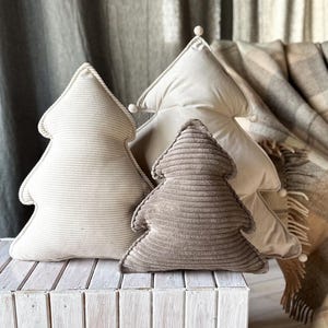 May include: Three tree-shaped decorative pillows in varying sizes and shades of beige and brown. The pillows are made of corduroy fabric and are arranged on a white wooden surface. A plaid blanket is in the background.
