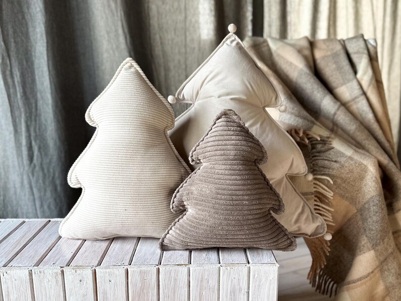 May include: Three tree-shaped decorative pillows in varying sizes and shades of beige and brown. The pillows are made of corduroy fabric and are arranged on a white wooden surface. A plaid blanket is in the background.