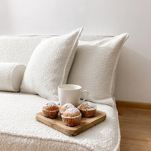 May include: A cozy scene featuring a white textured sofa with matching pillows. A wooden tray holds four muffins dusted with powdered sugar, alongside a white mug. The setting suggests a comfortable and inviting space.
