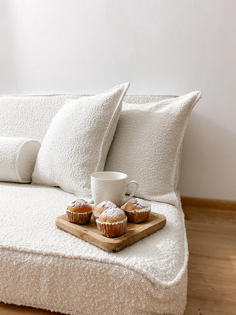 May include: A cozy scene featuring a white textured sofa with matching pillows. A wooden tray holds four muffins dusted with powdered sugar, alongside a white mug. The setting suggests a comfortable and inviting space.