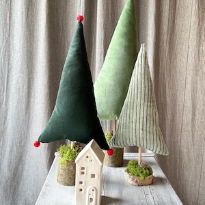 May include: A collection of decorative Christmas trees in varying shades of green, with red pom-poms and wooden bases. A white ceramic house with cut-out windows sits in front of the trees on a distressed white wooden table.