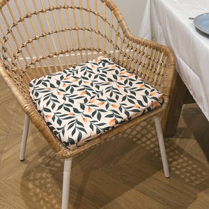 May include: A square cushion with a botanical pattern of green leaves and orange fruit on a light pink background rests on a wicker chair with white legs. The chair is positioned near a table with a white tablecloth.