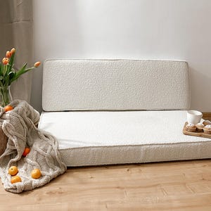 May include: A cozy, off-white, textured sofa with a matching backrest. A neutral-toned cable-knit throw blanket, fresh orange tulips in a glass vase, and a wooden tray with muffins and a cup complete the scene. The sofa is a comfortable seating option.