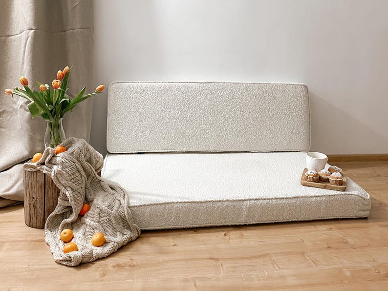 May include: A cozy, off-white, textured sofa with a matching backrest. A neutral-toned cable-knit throw blanket, fresh orange tulips in a glass vase, and a wooden tray with muffins and a cup complete the scene. The sofa is a comfortable seating option.