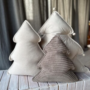 May include: Three Christmas tree-shaped decorative pillows in varying shades of beige and brown. The pillows are made of corduroy fabric and are arranged on a white wooden surface. The background features a gray curtain.