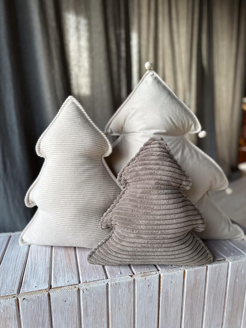 May include: Three Christmas tree-shaped decorative pillows in varying shades of beige and brown. The pillows are made of corduroy fabric and are arranged on a white wooden surface. The background features a gray curtain.