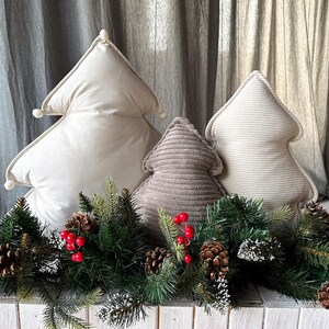 May include: Three decorative Christmas tree-shaped pillows in cream and taupe hues. The pillows are arranged in front of a festive garland with pinecones and red berries. The pillows add a touch of holiday cheer.