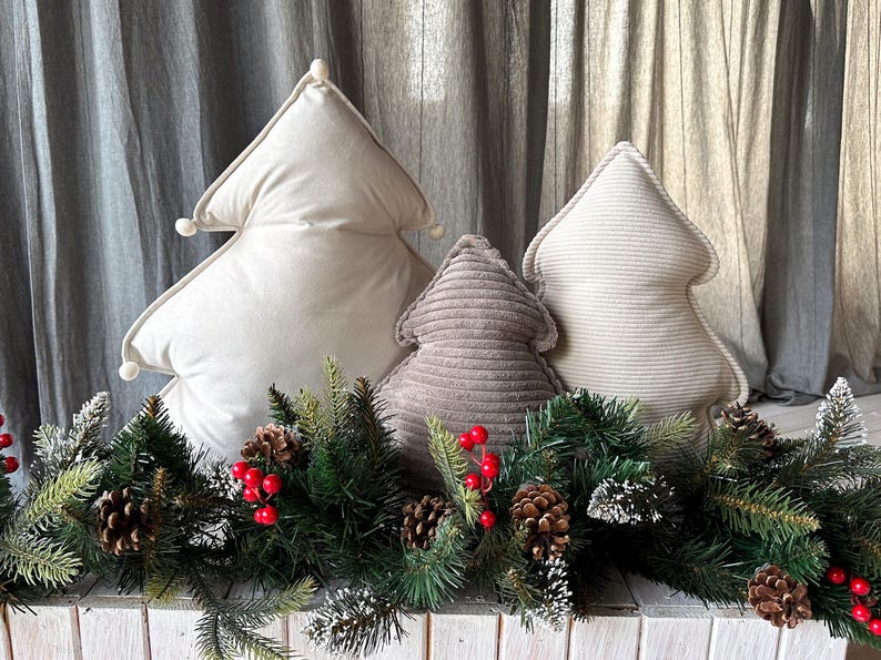 May include: Three decorative Christmas tree-shaped pillows in cream and taupe hues. The pillows are arranged in front of a festive garland with pinecones and red berries. The pillows add a touch of holiday cheer.