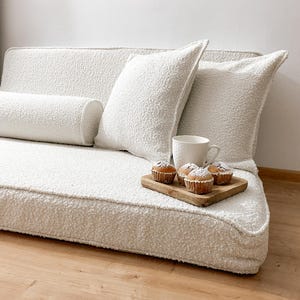 May include: A cozy, off-white boucle sofa with matching pillows and a cylindrical bolster. A small wooden tray holds muffins and a white mug. The sofa sits on a light wood floor, creating a warm, inviting scene.
