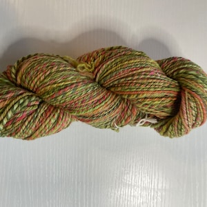 May include: A skein of hand-dyed yarn in shades of green, red, and yellow. The yarn is tightly wound and appears soft, ideal for knitting or crocheting projects. The colors blend together, creating a variegated effect.