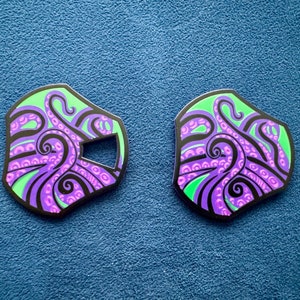 Puede incluir: Dos pines de metal negro con un diseño de pulpo verde y morado. Los tentáculos del pulpo están delineados en negro y tienen un patrón de remolino.