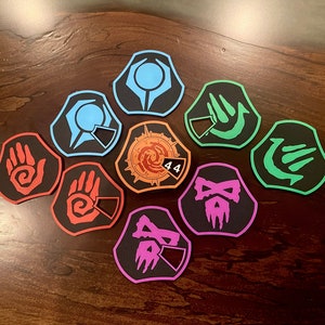 May include: Nine colorful, octagonal game tokens with different symbols and a number "44" on one of the tokens. The tokens are on a wooden surface.