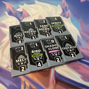May include: A collection of seven rectangular game tokens with grey borders and black centers. Each token features white text including labels like Gold, Recruit, Sand Soldier, Sprite, Mech, Bird, Shadow Clone, and Tentacle, along with numerical values.