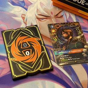 Peut inclure: Un compteur de jeu rectangulaire noir et orange avec un cadran central affichant le chiffre zéro, placé à côté d'une carte à collectionner rectangulaire illustrant une personne avec le texte Defender of Tomorrow.