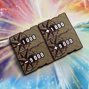 Puede incluir: Cuatro fichas de juego cuadradas de color verde oliva con un diseño de rosa de los vientos en marrón oscuro. Cada ficha muestra un valor numérico en texto blanco: -1000, +1000, +9000 y -9000. Las fichas tienen un borde dentado y acabado mate.