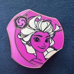 Puede incluir: Un pin de esmalte rosa y blanco con una ilustración estilizada de una mujer con cabello largo blanco y rostro morado. El pin tiene un diseño de espiral en blanco y negro en la esquina superior izquierda.