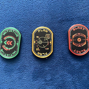 Puede incluir: Tres fichas redondas de plástico negro con bordes y texto de diferentes colores. La primera ficha tiene un borde verde y el texto "INITIATIVE" escrito alrededor del borde. La segunda ficha tiene un borde amarillo y el texto "PLAN" escrito alrededor del borde. La tercera ficha tiene un borde rojo y el texto "BLAST" escrito alrededor del borde.