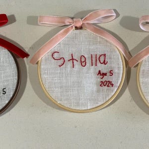May include: Three embroidered hoop ornaments with pink and red ribbon. The ornaments are personalized with the names "Brynn", "Stella", and "Lily" and their ages.