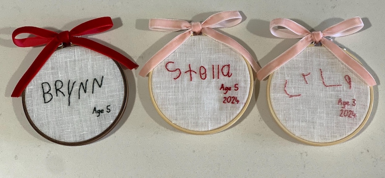 May include: Three embroidered hoop ornaments with pink and red ribbon. The ornaments are personalized with the names "Brynn", "Stella", and "Lily" and their ages.