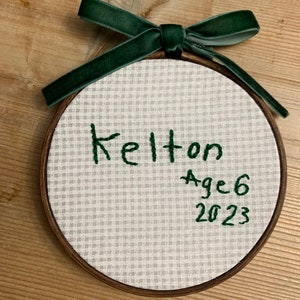May include: A white fabric ornament with a green embroidered ribbon and the text "Kelton Age 6 2023".