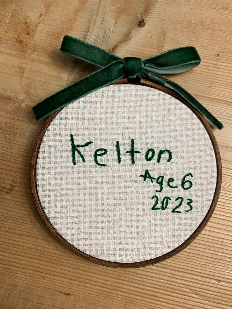 May include: A white fabric ornament with a green embroidered ribbon and the text "Kelton Age 6 2023".