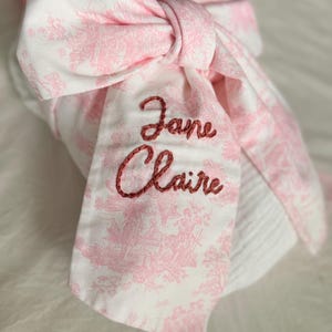 May include: A pink and white patterned bow with the embroidered names "Jane Claire" in a cursive font. The bow is made of fabric with a floral design and is tied in a large knot.
