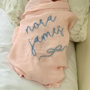 May include: A light pink baby swaddle blanket with the name "Nora James" embroidered in blue script. A blue bow design with pearl accents is embroidered near the bottom. The blanket is wrapped around a baby.