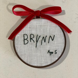 May include: A white fabric circle with a brown wooden frame, embroidered with the name "BRYNN" and the text "Age 5". A red velvet ribbon is tied around the top of the frame.