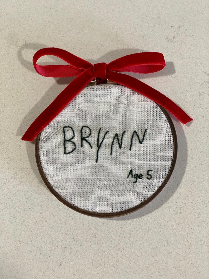 May include: A white fabric circle with a brown wooden frame, embroidered with the name "BRYNN" and the text "Age 5". A red velvet ribbon is tied around the top of the frame.