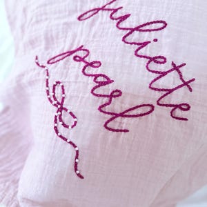 Könnte beinhalten: Hellrosa Stoff mit dem gestickten Namen "Juliette Pearl" in Schreibschrift. Die Stickerei ist in einem tiefen Magenta gehalten, mit einem dekorativen Perlenakzent.