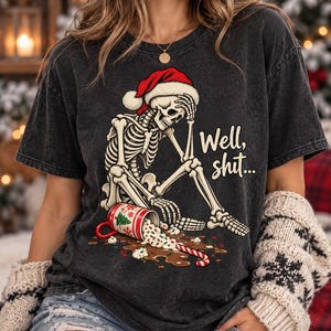 May include: A charcoal gray t-shirt featuring a graphic of a skeleton wearing a red Santa hat, sitting with its head in its hand. The text reads Well, shit... next to a spilled mug of hot cocoa with a candy cane and green sprinkles.