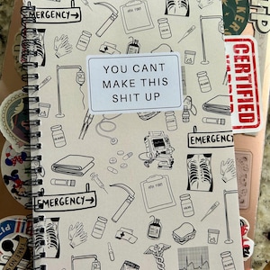 Puede incluir: Cuaderno de espiral con temática médica. La portada presenta ilustraciones de equipos médicos y el texto "YOU CANT MAKE THIS SHIT UP." El cuaderno está rodeado de pegatinas variadas.