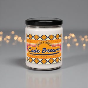 May include: A white candle in a glass jar with a black lid. The label features a colourful geometric design in orange, yellow, and blue, with the text "Smells Like Code Brown" in a stylized font. The candle is on a grey surface.