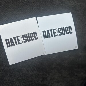 May include: Two white square stickers with black text that reads "DATE BEFORE SUCC" on a black fabric background.