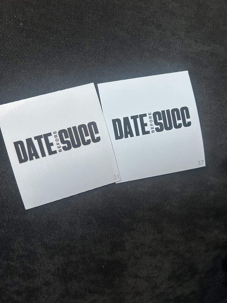 May include: Two white square stickers with black text that reads "DATE BEFORE SUCC" on a black fabric background.