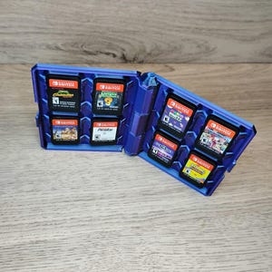 以下が含まれることがあります： Nintendo Switchのゲームカートリッジを収納する、青色のヒンジ付きケース。 開いたケースには、カラフルなラベルが付いた複数のゲームカートリッジが表示されています。 ケースは明るい色の木製の表面に置かれています。