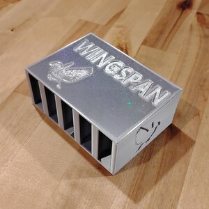 May include: A silver rectangular box with the word "WINGSPAN" engraved on the top. The box has multiple vertical slots and a bird design. The box is sitting on a wooden surface.