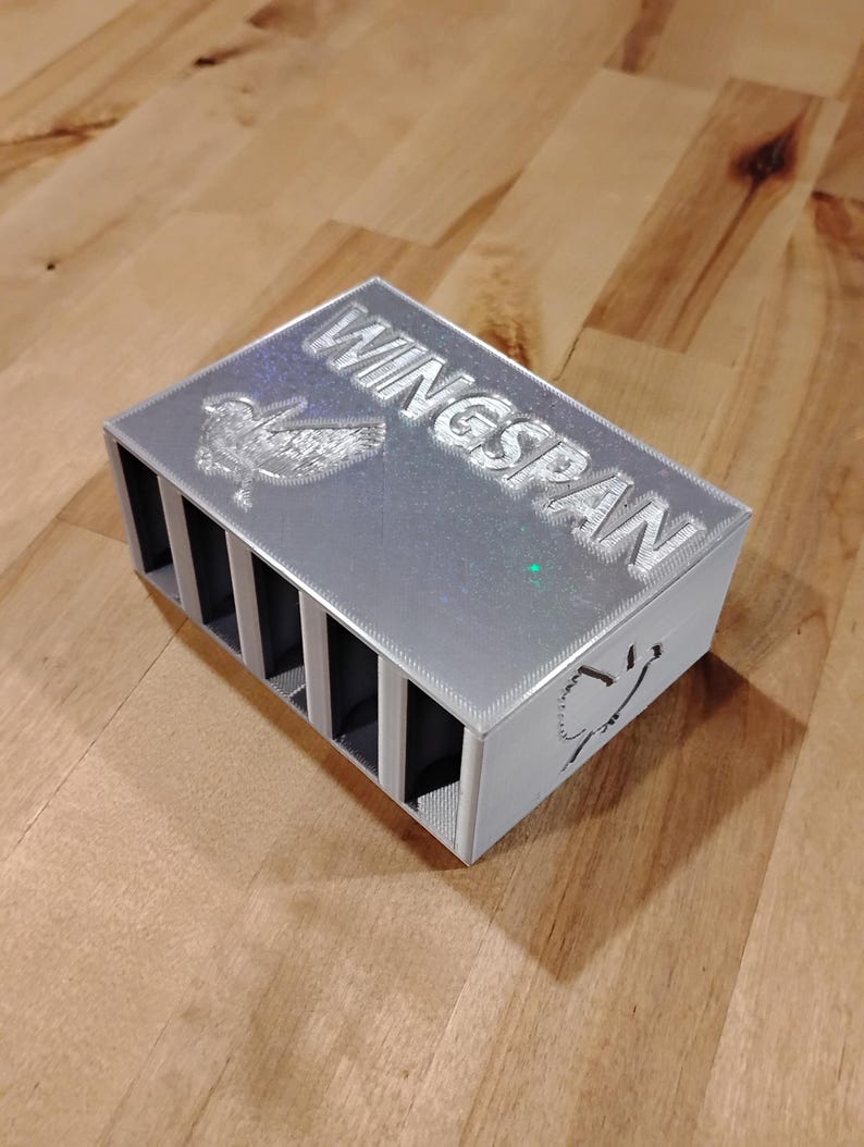 May include: A silver rectangular box with the word "WINGSPAN" engraved on the top. The box has multiple vertical slots and a bird design. The box is sitting on a wooden surface.