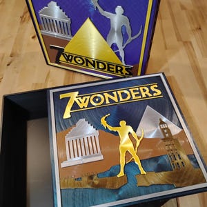 May include: Two-piece box set with the title "7 Wonders" in gold lettering. The lid features a gold pyramid, a silver figure, and architectural elements. The box has a blue and brown background with a silver border.