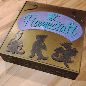 May include: A square, wooden box with the word "Flamecraft" in teal script on the top. The box has a gold-colored top with a purple oval and three dark gray dragon silhouettes. The box is on a wooden surface.