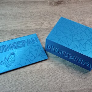 May include: Two-piece set of turquoise blue game storage boxes. The lid and the base of the larger box and the lid of the smaller box feature the word "WINGSPAN" and a bird graphic. Both boxes have a geometric pattern.